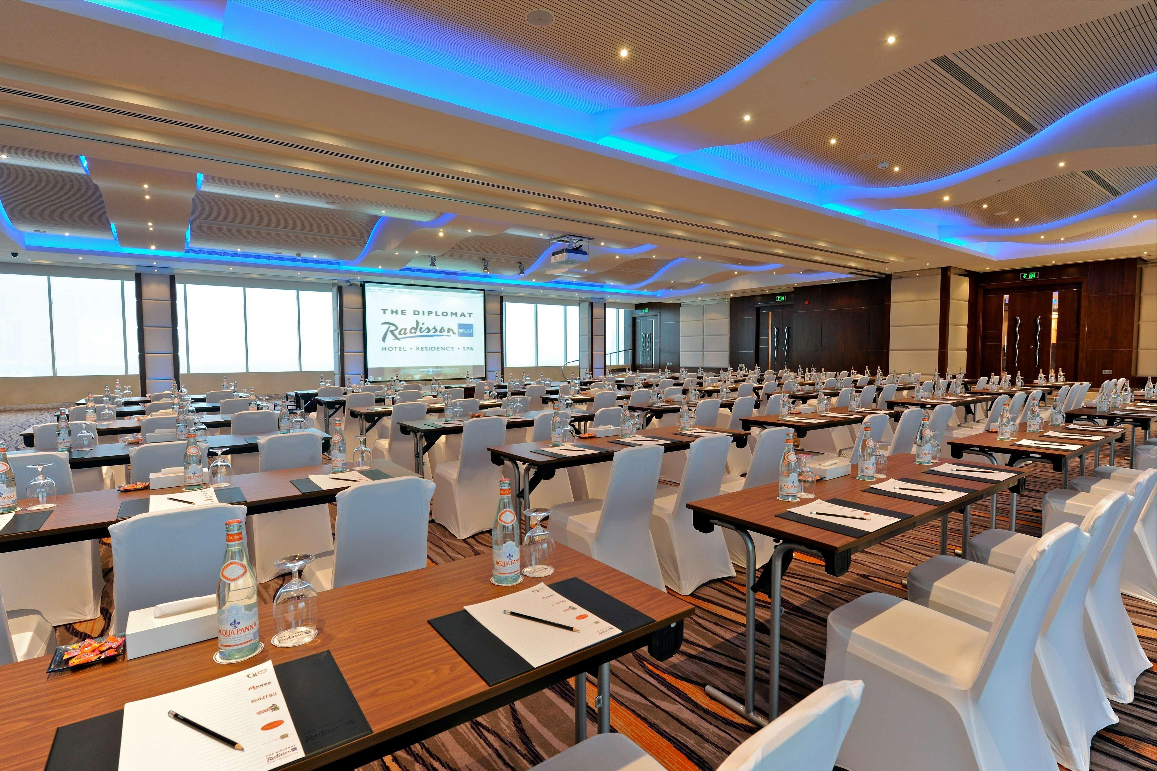 The Diplomat Radisson Blu Hotel Residence & Spa Manama Exterior photo The photo shows a spacious conference room set up for a meeting or event. Multiple rows of tables and chairs are arranged neatly, facing a large screen at the front. Each table has water bottles, notebooks, and pens provided for attendees. The room f