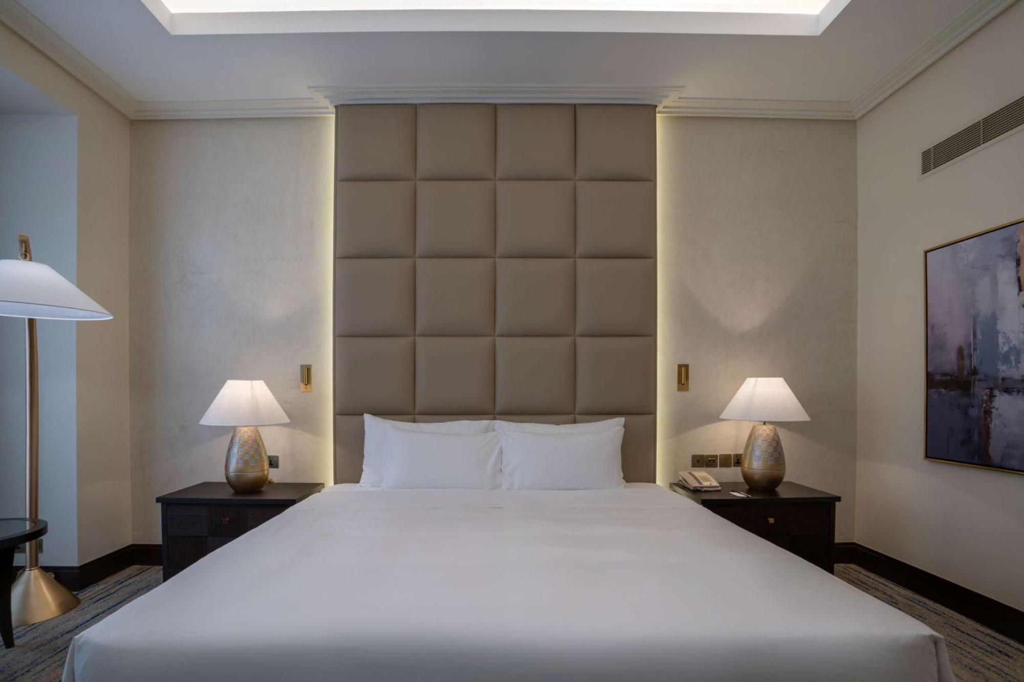 The Diplomat Radisson Blu Hotel Residence & Spa Manama Exterior photo The image shows a modern and elegantly designed hotel bedroom. The focal point is a large bed with a simple white comforter and two white pillows. Behind the bed is a padded headboard in a neutral color, which adds a touch of sophistication. On eithe