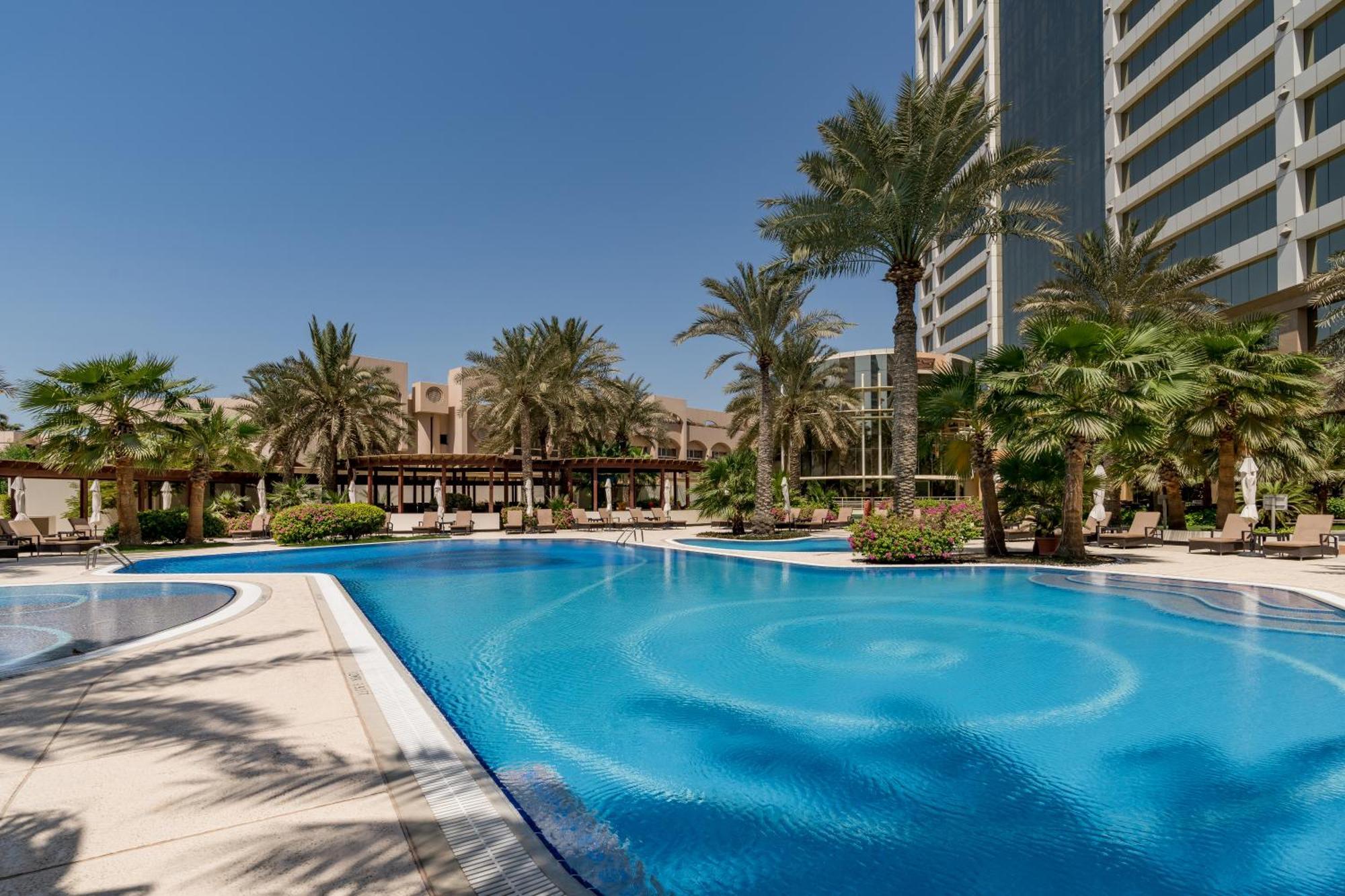 The Diplomat Radisson Blu Hotel Residence & Spa Manama Exterior photo The photo shows a large, inviting swimming pool surrounded by lush palm trees and greenery. The pool has a beautiful blue color and features a circular design pattern in the water. In the background, there are tall buildings, suggesting a resort or h