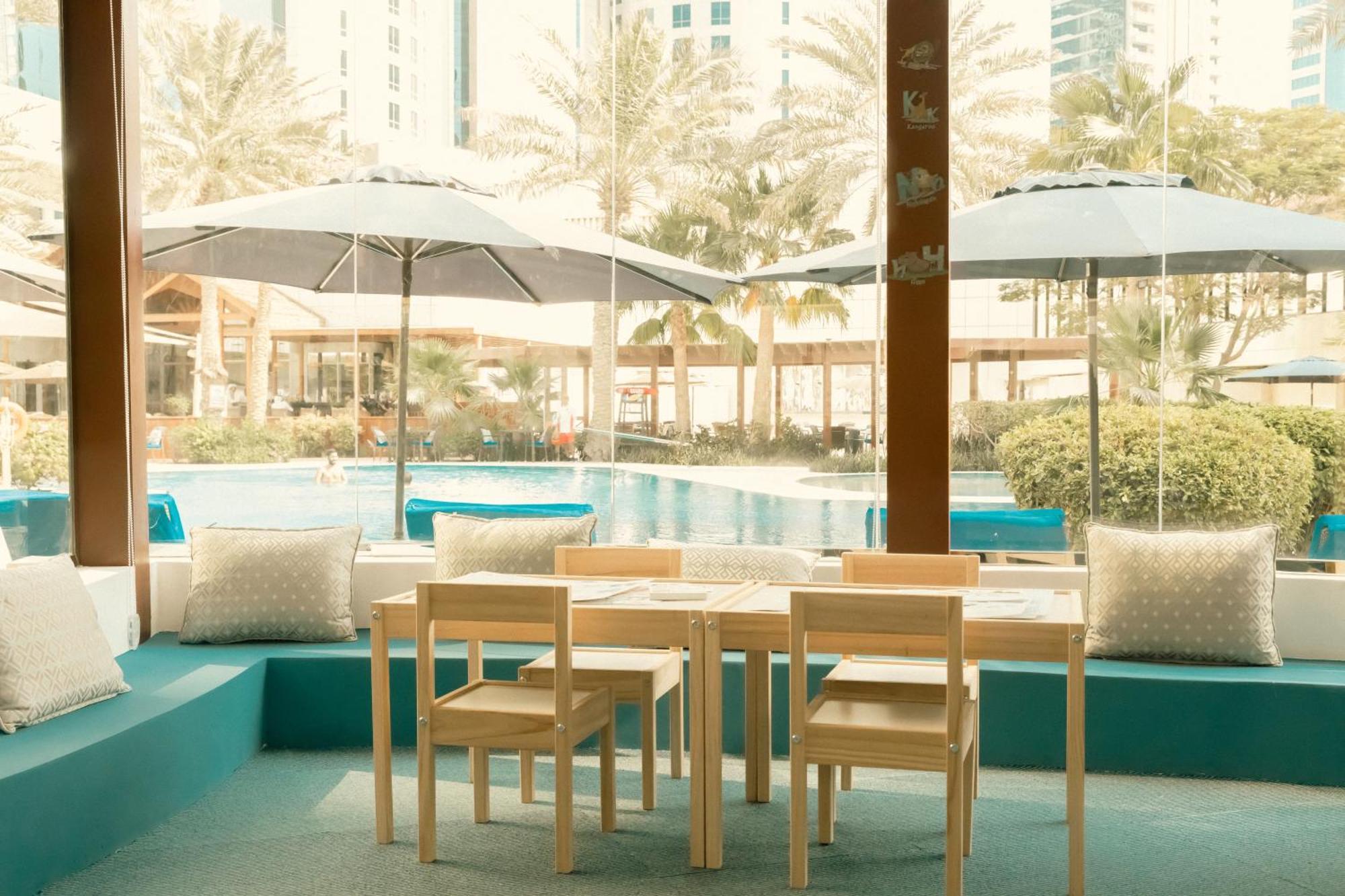 The Diplomat Radisson Blu Hotel Residence & Spa Manama Exterior photo The photo depicts a cozy indoor seating area with a small wooden table and four wooden chairs. Behind the seating area, large windows provide a view of an outdoor space featuring a swimming pool, palm trees, and umbrellas for shade. The color scheme 