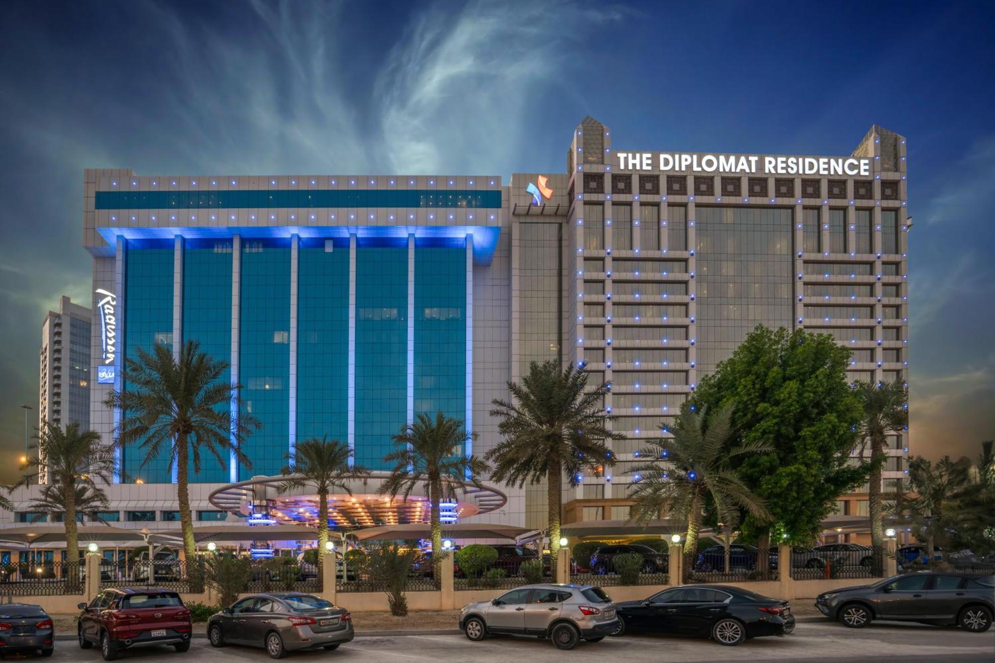 The Diplomat Radisson Blu Hotel Residence & Spa Manama Exterior photo The photo features two large buildings, likely hotels or residences. On the left, there’s a blue glass facade with illuminated signage, possibly indicating a hotel or establishment, with trees in front. On the right, there’s a building labeled "The D