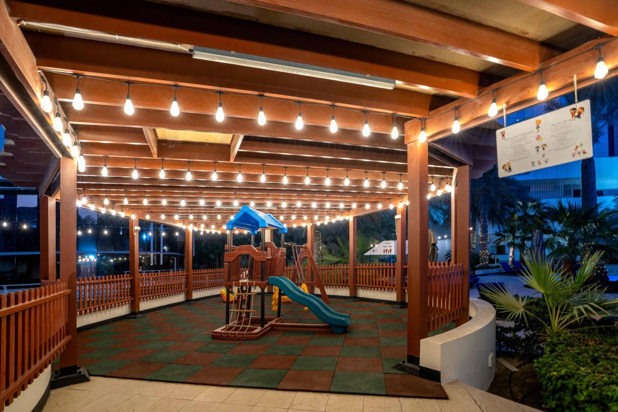 The Diplomat Radisson Blu Hotel Residence & Spa Manama Exterior photo The photo shows a playground area that's covered by a wooden structure. The playground features a colorful playset, including a slide and climbing elements. It's surrounded by a safety mat and is illuminated by string lights hanging overhead, creatin